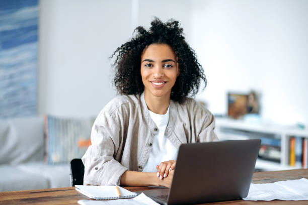 schöne freundliche, selbstbewusste junge afroamerikanerin in stilvoller freizeitkleidung, freiberuflerin, managerin oder studentin, die am laptop sitzt, zu hause arbeitet oder studiert, in die kamera schaut, lächelt - studentin stock-fotos und bilder