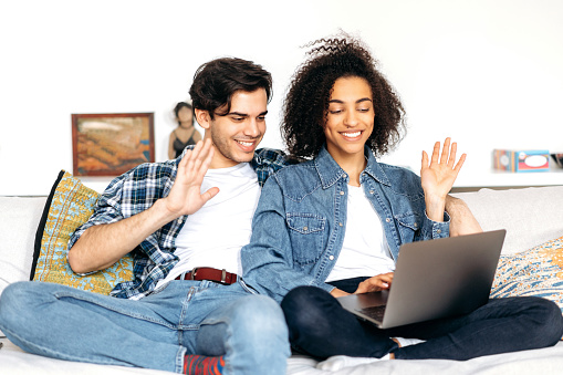 Happy young multiracial couple spends leisure time at home on the sofa, using a laptop, they talking via video conference with family or friends, greeting interlocutor, smiling, happy together