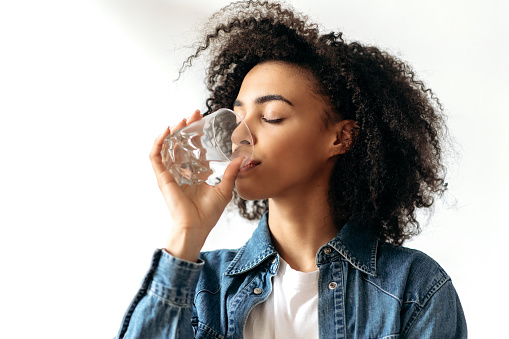 Healthy lifestyle. African american curly young woman drinks with pleasure a glass of pure water. Beautiful healthy woman follow healthy lifestyle, cares about health