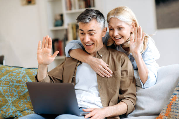 istock Happy caucasian joyful spouses spend time at home on a sofa, use laptop for online communication with friends or family by video conference, greeting by waving hands, smiling. Video call concept 1334906191