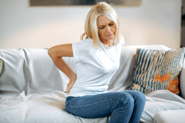 腰痛に苦しむ動揺した成熟した女性、リビングルームのソファに座っている不幸なシニアブロンド、背中の痛みのために不快感を感じ、関節の問題を抱えている、休息が必要 - arthritis osteoporosis pain backache ストックフォトと画像