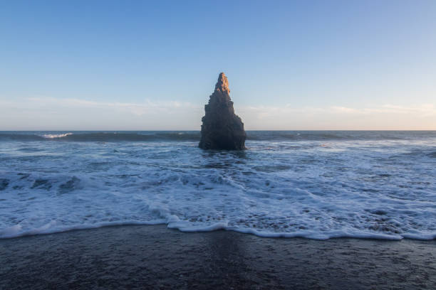 istock Tall pointy rock on beach shoreline with waves rolling in over sand 1334906336