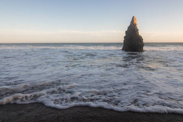 istock Tall pointy rock on beach shoreline with waves rolling in over sand 1334906393