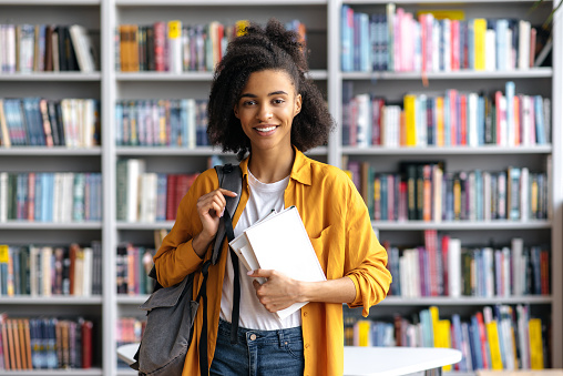 Portrait of an african american happy pretty, smart confident female student with backpack, stylishly dressed, holding books, stands in the university library, looks at the camera, smiling friendly