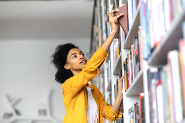 studentessa afroamericana si trova nella biblioteca universitaria, alla ricerca di un libro. una ragazza piuttosto intelligente che cerca informazioni per un progetto, scegliendo il libro alla libreria della biblioteca, il concetto di educazione - book library bookshelf university foto e immagini stock