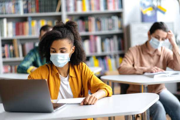 istock Young students study during pandemic, sitting in university at distance from each other wearing protective medical masks. African american female student taking notes during lecture, using laptop 1334911411