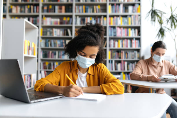 istock Multiethnic students study during the pandemic, sit at a tables at distance in a protective medical masks in the library with a laptop, listen to lecture, African American girl takes notes 1334911417