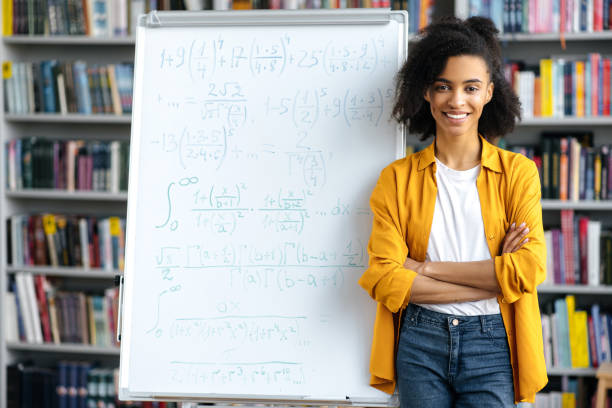 portret uroczej radosnej dziewczyny, afroamerykańskiej nauczycielki, stoi przy tablicy, ubrana w zwykłe ubrania, prowadzi lekcję, krzyżuje ręce, patrzy w kamerę, uśmiecha się przyjaźnie - education classroom advice mathematics zdjęcia i obrazy z banku zdjęć