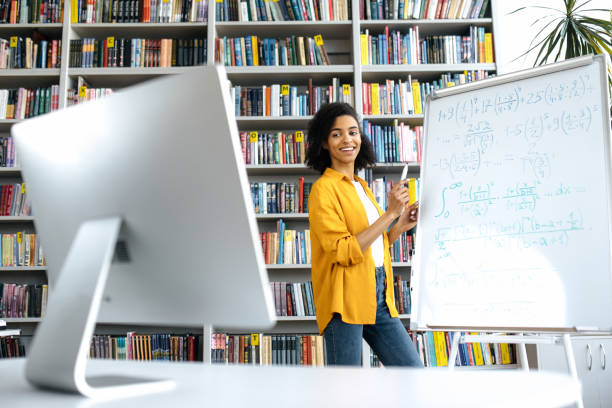 Pleasant african american female teacher conducts online lesson for students via video conference by computer, stands at university library, shows information on whiteboard, looks at computer, smile la agradable maestra afroamericana lleva a cabo una lección en línea para los estudiantes a través de una videoconferencia por computadora, se encuentra en la biblioteca de la universidad, muestra información en la pizarra, mira la computadora, sonríe