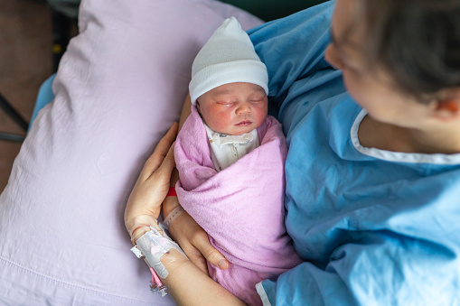 High angle view looking down at an adorable mixed race Asian newborn baby sleeping in her mother's arms at the hospital right after birth. The healthy and tiny infant is swaddled in a pink baby blanket and is wearing a white knit hat. Family, love and bonding concepts.