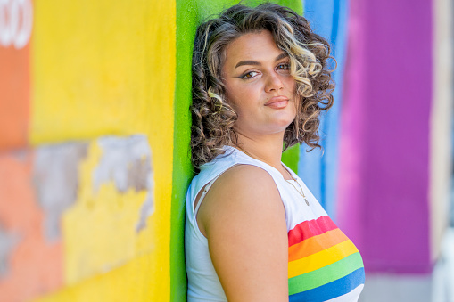 A beautiful young female adult of Latin descent standing outdoors on a bright day. She wears a sleeveless white top with the Pride (rainbow) colours on it. She is leaning against a pride mural wall. She is happy and smiling.