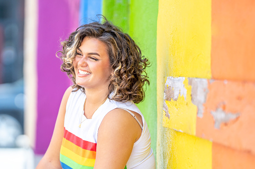 A beautiful young woman of Latin descent standing outdoors on a bright day. She wears a sleeveless white top with the Pride (rainbow) colours on it. She is sitting in front of a pride mural. She is happy and smiling.