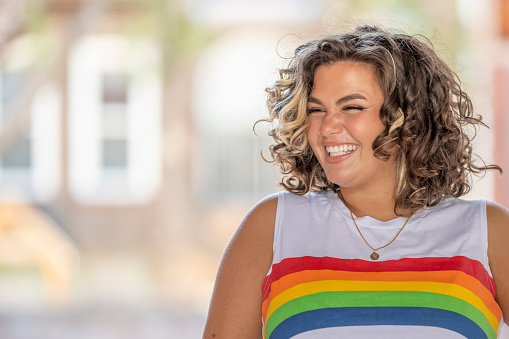 A beautiful young woman of Latin descent standing outdoors on a bright day. She wears a sleeveless white top with the Pride (rainbow) colours on it. She is happy and smiling.