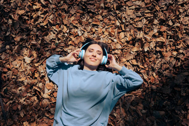 mujer sentada en hojas secas de otoño escuchando música - orange white audio fotografías e imágenes de stock