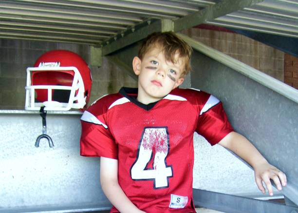 retrato de un niño pequeño con uniforme de fútbol sentado debajo de las gradas - foto de archivo - youth league american football childhood helmet fotografías e imágenes de stock