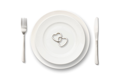 Overhead shot of white dinner plate with two silver heart shaped rings and cutlery on white background.