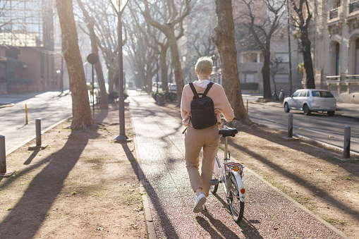 A young millennial woman walking down the city street, carrying a bicycle in her hands. Concept of healthy life. Ecological mode of transportation. Horizontal image. Copy space for text.