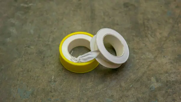 White tape or thread seal tape with yellow casing on the floor. It is a polytetrafluoroethylene (PTFE) film tape commonly used in plumbing for sealing pipe threads. Selective focus on object.