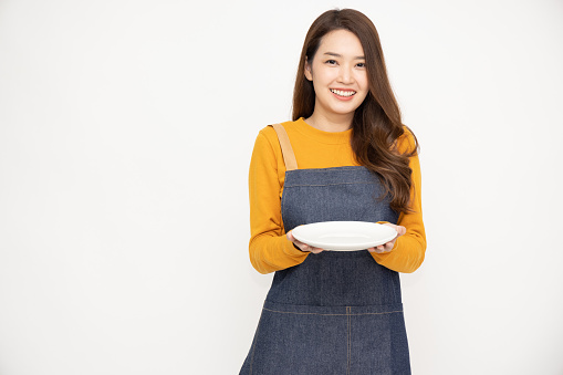 Young Asian woman in apron standing and holding empty white plate or dish isolated on white background
