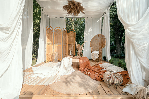 Garden arbour with rustic beige decoration, outdoor design. Wicker furniture outside in boho pergola, modern home backyard. Summer arbor with nature plant, wooden decor and blankets.