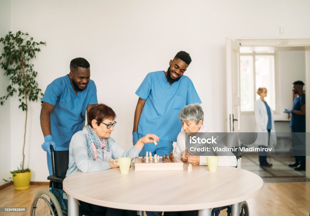 Two older women playing a game of chess, overseen by two male nurses Two older  women playing  a game of chess, overseen by two male nurses.  In a common room in a hopsital, in the background a doctor and assistant talk. African Ethnicity Stock Photo
