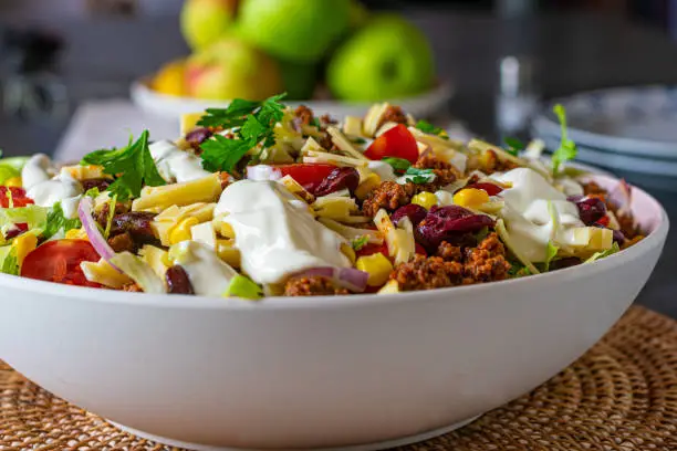 A large bowl of fresh cooked taco salad with ground beef, kidney beans, lettuce and vegetables. Topped with sour cream and served on a table. Isolated and front view
