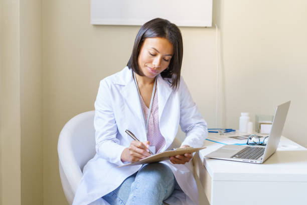 Young african woman doctor in white coat sitting at workplace in clinic and filling patient form stock photo