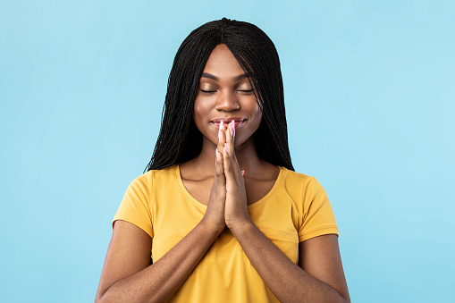 Happy African American Millennial Lady Praying With Eyes Closed Making Prayer's Gesture Standing Over Blue Background. Studio Shot Of Peaceful Black Female In Pray Concept