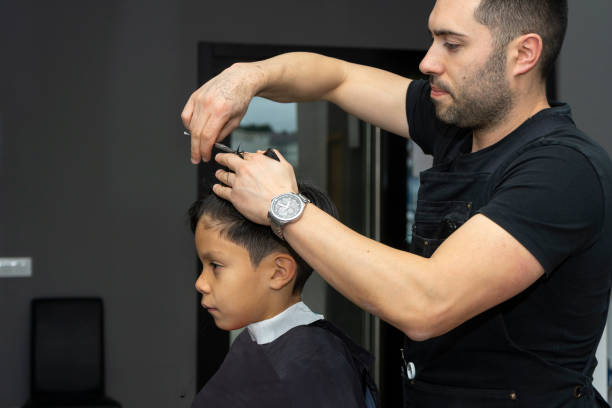 Barber cuts boy's hair with scissors while he is calmly looking at himself in mirror stock photo