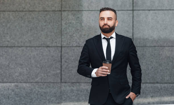 istock Confident middle-aged businessman in suit with takeaway coffee standing outdoors against grey wall, copy space 1334994622