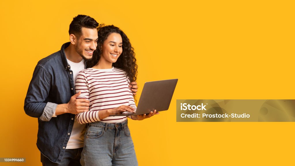 Young middle eastern couple using laptop together while standing over yellow background Young middle eastern couple using laptop together while standing over yellow background in studio, cheerful arab spouses shopping online or browsing new website on computer, panorama with copy space Couple - Relationship Stock Photo