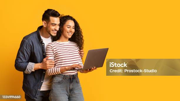Young Middle Eastern Couple Using Laptop Together While Standing Over Yellow Background Stock Photo - Download Image Now