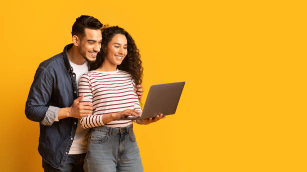 jovem casal do oriente médio usando laptop juntos enquanto estava em pé sobre fundo amarelo - sale relationships loving indoors - fotografias e filmes do acervo