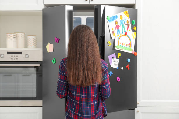 frau, die die kühlschranktür mit kinderzeichnungen, notizen und magneten in der küche öffnet - refrigerator domestic kitchen magnet door stock-fotos und bilder