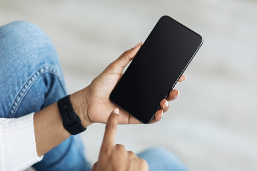 Mockup, blank screen of smartphone. Lady hand texting, using cellphone on sofa at home. Young african american woman typing on smartphone with empty screen, closeup