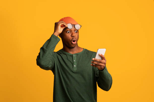 increíble aplicación. tipo afroamericano sorprendido mirando la pantalla del teléfono inteligente - sorpresa fotografías e imágenes de stock