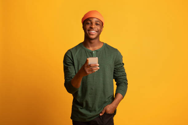 オンラインコミュニケーション。スマートフォンを持ち、カメラに微笑む陽気な若い黒人男性 - ヒップスター ストックフォトと画像