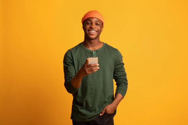 Online Communication. Cheerful young black man holding smartphone and smiling at camera Online Communication. Portrait of cheerful young black man holding smartphone and smiling at camera, happy millennial african guy using cellphone while standing over yellow background, copy space hipsters stock pictures, royalty-free photos & images