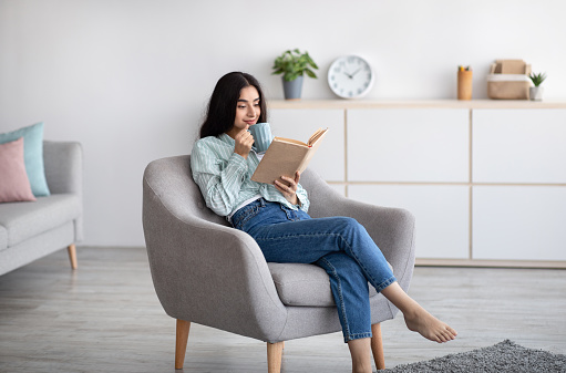 Attractive Indian lady with cup of aromatic coffee and open book relaxing in armchair at home. Pretty millennial woman enjoying hot beverage and reading exciting story, having lazy weekend indoors