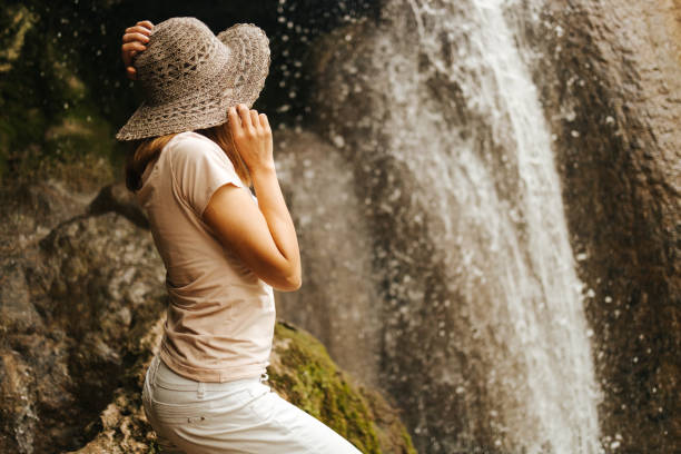 istock Beautiful young woman with a hat on her head standing next to a waterfall 1335002780