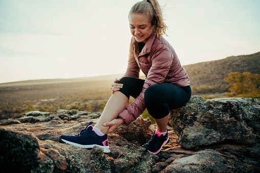 Caucasian female scrunching face hiking on mountain holding sprained ankle in pain waiting for help sitting on rock at sunset. High quality photo