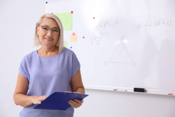 istock Mature teacher with clipboard near whiteboard in modern classroom. Space for text 1335010695