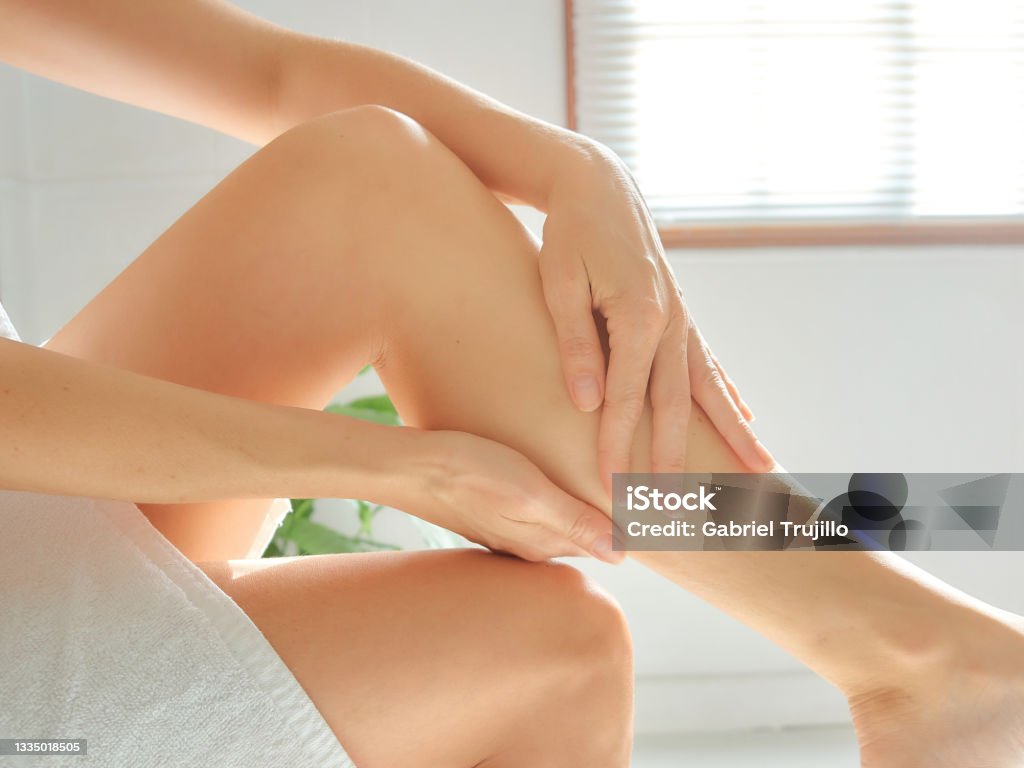 Young Caucasian woman depilates her leg with an electric epilator device in the bathroom. Wellness and skin care concept. Young woman massaging her legs after depilation in the bathroom. Massage Oil Stock Photo