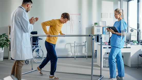 Hospital Physical Therapy Room: Patient with Injury Walking Holding for Parallel Bars, Professional Physiotherapist and Doctor Assist, Help, Train Disabled Person Do Rehabilitative Physiotherapy