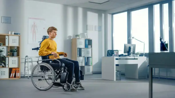 Photo of Hospital Physical Therapy: Determined Young Man with Leg Injury is Sitting on a Wheelchair. Miracle of Rehabilitation, Strong Mindedness, Willpower, Professional Doctors.