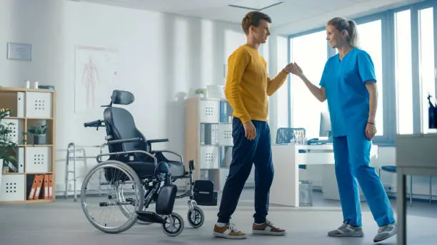 Photo of Hospital Physical Therapy: Determined Young Man with Injury Successfully Stands up from Wheelchair and Walks with Help of Rehabilitation Physiotherapist. Willpower, Grit, Strong Mindedness.