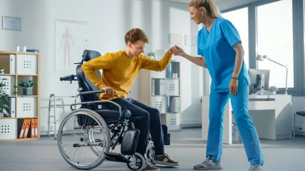 Photo of Hospital Physical Therapy: Determined Young Man with Injury Successfully Stands up from Wheelchair and Walks with Help of Rehabilitation Physiotherapist. Willpower, Grit, Strong Mindedness.