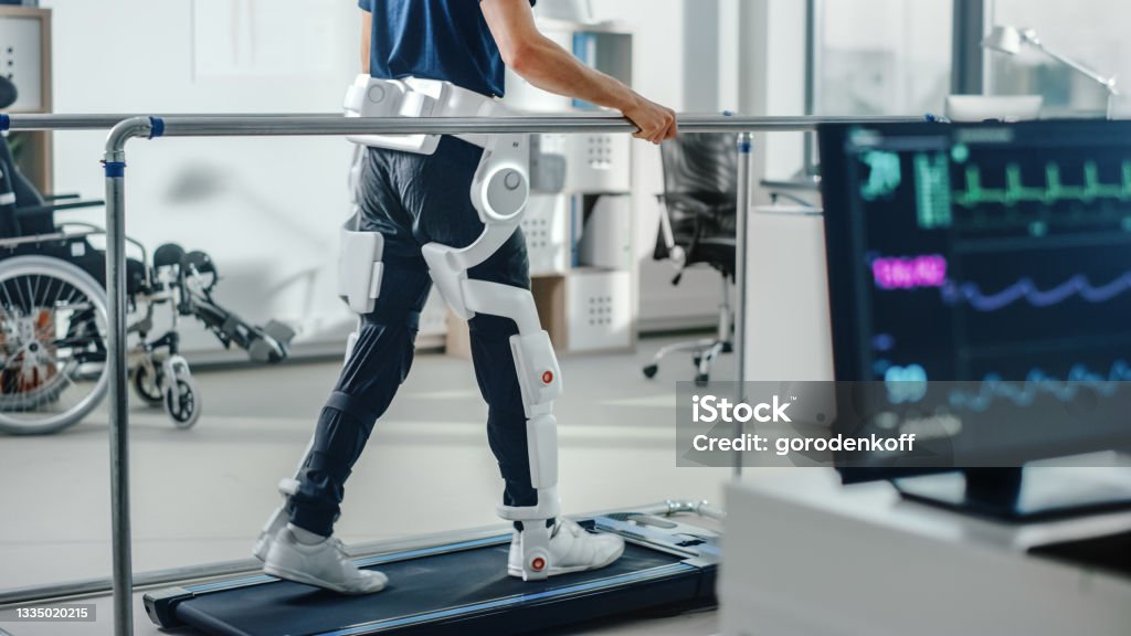 Moderne Krankenhaus-Physiotherapie: Patient mit Verletzungen geht auf dem Laufband mit fortschrittlichen Roboter-Exoskelett-Beinen. Physiotherapie Rehabilitationstechnologie, um behinderte Menschen laufen zu lassen. Fokus auf Beine - Lizenzfrei Gesundheitswesen und Medizin Stock-Foto