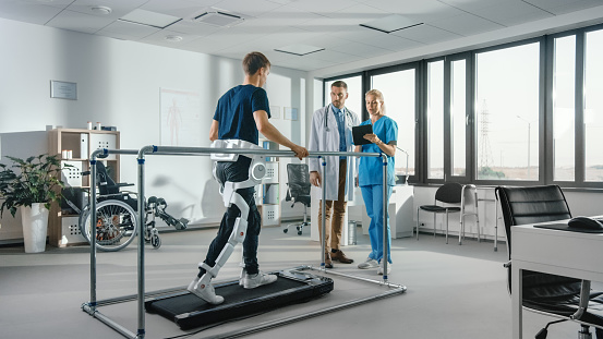 Modern Hospital Physical Therapy: Patient with Injury Walks on Treadmill Wearing Advanced Robotic Exoskeleton. Physiotherapy Rehabilitation Scientists, Engineers, Doctors use Tablet Computer to Help