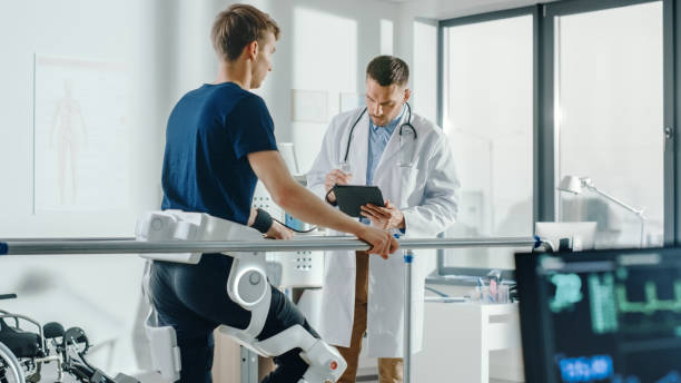 istock Modern Hospital Physical Therapy: Doctor Uses Tablet Computer, Helps Disabled Patient with Injury Walk on Treadmill Wearing Advanced Robotic Exoskeleton Legs. Physiotherapy Rehabilitation Technology 1335020218